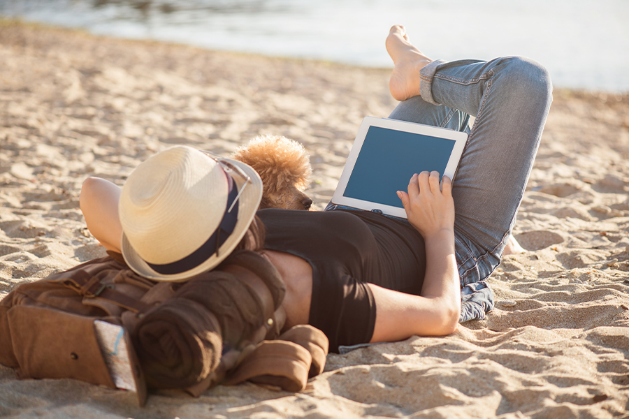 Client Center - Woman Traveler with Her Dog Holding Tablet While Relaxing Outdoors During on the Beach